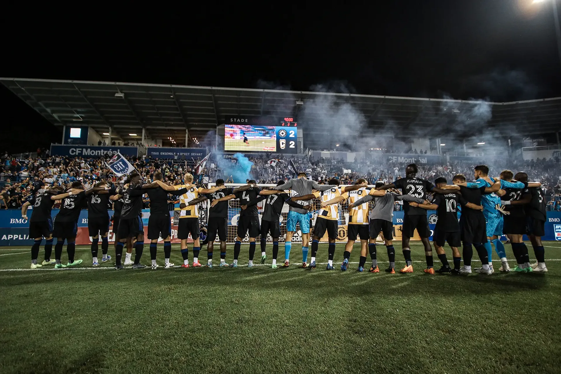 CF Montréal players arm-in-arm facing a packed Stade Saputo as blue and white smoke rises from the supporter section